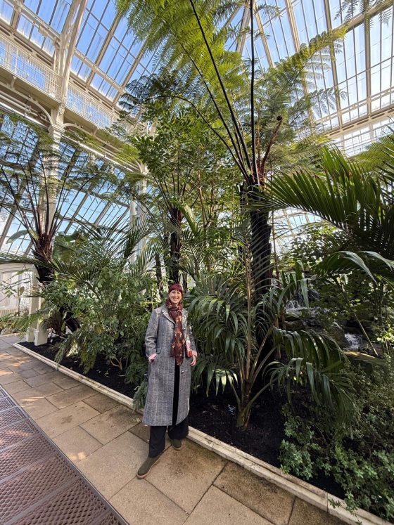 A person in a long gray coat, standing inside a conservatory featuring tall palms. 